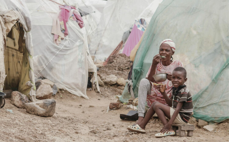 Une mère et son fils africains démunis mangeant seuls devant un tente faite avec des matériaux de récupération dans un environnement humide
